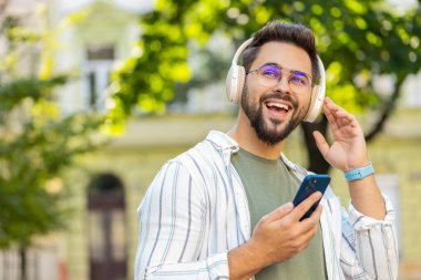 Kablosuz kulaklık takan mutlu beyaz adam dışarıda akıllı telefondan en sevilen enerjik müziği dinliyor. Şehir caddesinde yürüyen gülümseyen bir turist. Kasaba yaşam tarzları.