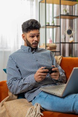 Dizüstü bilgisayarda video oyunu oynayan heyecanlı Hintli Arap adam boş vaktini kanepede geçiriyor. Bilgisayar bilgisayarında joystick kontrolörüyle video oyunu oynayan mutlu İspanyol serbest yazar. Dikey