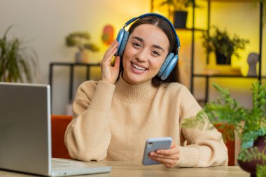 Happy relaxed overjoyed young woman in wireless headphones sits at home table looking at camera choosing listening favorite disco music in smartphone dancing entertaining. People weekend activities clipart