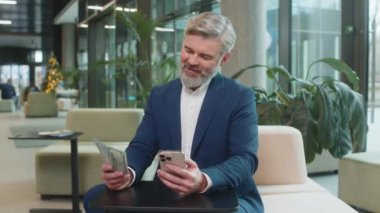 Başarılı, mutlu, olgun iş adamı akıllı telefon tutuyor ve ofis lobisinde oturmuş dolarları sayıyor. İnternette piyango kazanan beyaz sakallı adam. Bütçe hesaplanıyor.