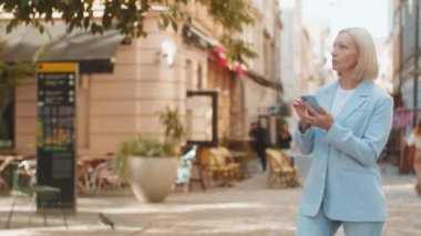 Resmi takım elbiseli beyaz iş kadını bir yön bulmak için mobil GPS kullanıyor. Şehir caddesinde yürürken mobil uygulamalarda yön arayan orta yaşlı kadın turist girişimcisi.