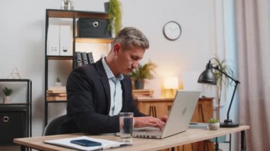 Kafkasyalı bir iş adamı, ofis laptopuyla çalışırken çalışma masasında oturup su içiyor. Takım elbiseli genç serbest çalışan bilgisayar ekranlı sörf internetine bakıyor