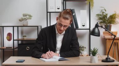 Genç iş adamının kameraya bakıp, iş arkadaşının, iş yerindeki görüntülü sohbete günlük yazmasını dinlediği görüntüler. Takım elbiseli şık adam masada notlar alıyor. Web kamerası görünümü.