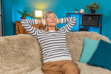 Beyaz, olgun bir kadın, evdeki kanepede rahatça oturuyor. Mutlu büyükanne gözlerini kapatır ve kablosuz kulaklıklarla sakin müzik dinlemeyi sever. Rahat ve huzurlu görünürken rahat bir odada dinlenir.