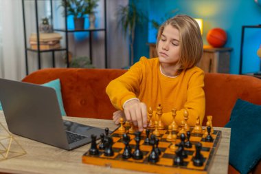 Beyaz çocuk evdeki laptop uygulamasında online satranç oynuyor. Masada taşları olan gerçek bir satranç tahtası kullanmak, düşünceli bir düşünce ve koltukta rahat bir şekilde otururken taşları hareket ettirmek.