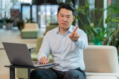 Portrait of mature businessman with laptop shakes finger and saying No be careful scolding and giving advice to avoid danger mistake disapproval sign sitting in lobby. Serious guy in shirt at hotel. clipart