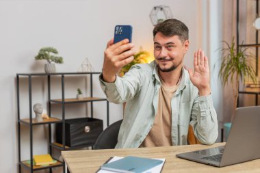 Kafkasyalı serbest çalışan genç adam sosyal medya için akıllı telefon görüntülerini kullanıyor. Masada oturmuş gülümsüyor ve ev ofisinden çalışıyor. Erkek öğrenci çalışması, e-öğrenme, uzaktan kumanda. Yaşam biçimi.