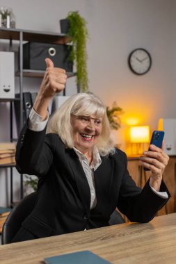 Mutlu olgun bir kadın akıllı telefondan yazı yazarak evet diyerek başarıyı kutluyor piyango kazanma hedefini başarıyor iyi haber zaferini oynuyor. Ofis masasındaki yaşlı kadın..