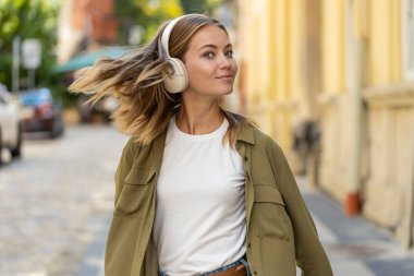 Kablosuz kulaklıklarla gezen mutlu, rahat genç kadın turistin arka görüntüsü açık havada dans eden favori enerjik disko müziğini dinliyor. Güneşli şehir sokağının önünden geçen beyaz bir genç kız..
