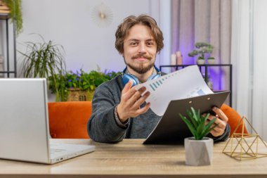 Mutlu gülümseyen adam evde belgeleri analiz ederek çalışıyor iş gazetelerini okuyor finansal raporları hazırlıyor. Beyaz adam laptoptaki ani zafer haberiyle şok oldu.