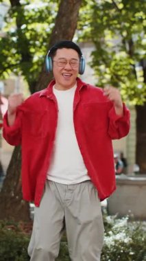 Aşırı mutlu Asyalı orta yaşlı adam kablosuz kulaklıkla dans ediyor favori enerjik müziği dinliyor, dışarıda zaferi kutluyor. Şehir sokağındaki Çinli adam. Kasaba yaşam tarzları. Dikey