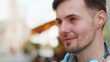 Kablosuz kulaklıklı, boynuna bakıp gülümseyen gündelik kıyafetlerle düşünceli genç bir turist. Şehir caddesinde duran mutlu beyaz erkek gezgin. Kasaba yaşam tarzları.