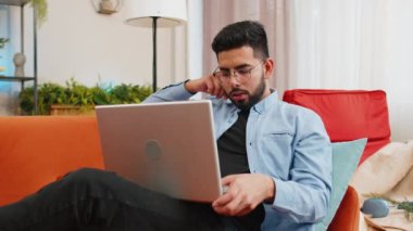 Laptop kullanan şaşırmış Hintli adamın yakın plan çekimi, iyi haberler almak ani zaferin şokuyla piyango ikramiyesini kutlamak. Netbook 'lu neşeli İspanyol adam evdeki oturma odasında kanepede oturuyor..