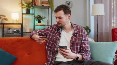 Ekose gömlekli üzgün genç adam elinde akıllı telefon ve kredi kartı kaşları çatık bir şekilde kanepede oturuyor ve internet üzerinden ödeme yapmaya çalışıyor. Parasızlık yüzünden kızgın hissediyor. Kafkasyalı dolandırıcılık kurbanı