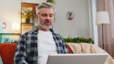 Gülümseyen sakallı, olgun bir adam dizüstü bilgisayarda yazı yazıyor. İnternette geziniyor. Kanepede tek başına oturan uygulamaları kullanıyor. Orta yaşlı, yaşlı, beyaz bir adam bilgisayarda çalışıyor. Oturma odasında ekrana bakıyor.