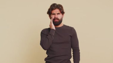 Indian bearded man touching sore cheek suffering from toothache cavities or gingivitis waiting for dentist appointment gums disease. Handsome Hindu guy indoors studio shot isolated on beige background
