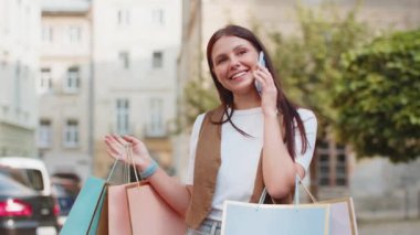 Alışveriş sonrası alışveriş torbaları açık havada satılan mutlu gülümseyen beyaz kadın. Kız turist şehir caddesinde durmuş akıllı telefondan uzaktan konuşma yapıyor..