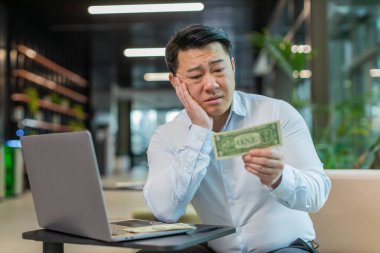 Depresyondaki Çinli işadamı maddi sorunlar hakkında endişeli. Tişörtlü stresli adam hayal kırıklığına uğramış görünüyor. Para borcu, kötü bir gün, bütçe kaybı, iflas modern ofis lobisinde oturuyor..