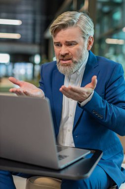 Takım elbiseli kızgın yetişkin patron, ofiste modern işyerinde otururken bilgisayarla video görüşmesi yapıyor. Tatminsiz iş adamı yöneticileri, şirketlerin sorunları hakkında astlarıyla iletişim kuruyor..