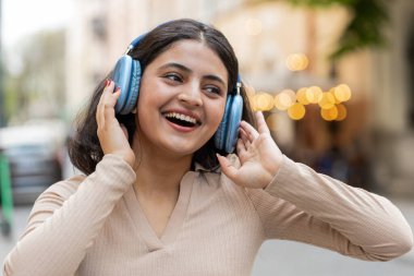 Kablosuz kulaklıklı mutlu Hintli genç kadın dışarıda akıllı telefondan dans ederek favori disko müziğini dinliyor. Şehir caddesinde yürüyen İspanyol kız. Kasaba yaşam tarzları