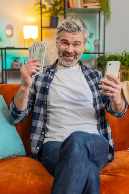 Başarılı, mutlu, son sınıf öğrencisi akıllı telefon tutuyor ve evdeki kanepede oturan banknotları sayıyor. İnternette piyango kazanan mutlu beyaz adam. Bütçe kavramı hesaplanıyor. Dikey