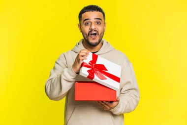 Mutlu Hintli genç adam kırmızı kurdeleli doğum günü hediyesi kutusunu açıyor. Tatil sürprizi konsepti. Gülümseyen Arap sakallı adam sarı stüdyo arka planında izole edilmiş büyük bir kutlama hediyesi aldı.