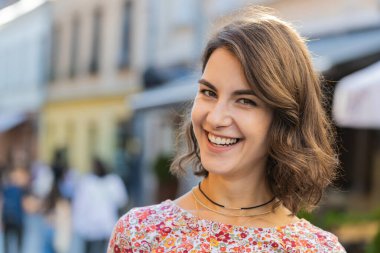 Mutlu güzel bir genç kadının portresi. Dostça gülümseyen, kameraya bakan mutlu yüz ifadesi. Hayal kuran, dinlenen, rahatlayan, dışarıda iyi haberler veren. Şehir güneşli yaz sokağında bir kız