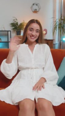 Kameraya gülümseyen, el sallayan, el sallayan, evdeki oturma odasında kanepede otururken selamlaşan mutlu beyaz kadın. Mutlu kız evde video kaydediyor. Dikey