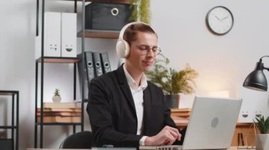 Mutlu genç iş adamı kablosuz kulaklık takıyor müzik dinlerken eğleniyor. Gülümseyen bir çalışan işte stres hissetmez. Resmi takım elbiseli neşeli beyaz adam ofis masasında dinlenmeyi sever..