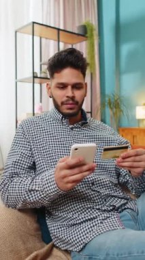 Hintli adam parayı transfer ederken kredi kartı ve akıllı telefon kullanıyor. Ev içinde alışveriş yaparken nakit para almıyor. Mutlu adam kanepede oturuyor. Dikey