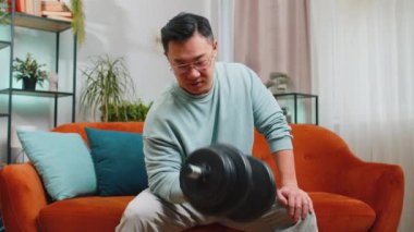 Asyalı adam halterleri kaldırıyor, bisepsi eğitiyor, kalori yakıyor, antrenman yapıyor, ev ortamında aerobik yapıyor. Sportif adam kanepede oturuyor.