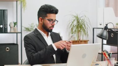 Rich Indian businessman working on laptop pc counting money cash, calculate earnings income profit at home office workplace. Professional manager freelancer man. Business people. Employment occupation