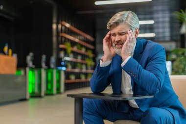 Baş ağrısı, tansiyon, migren ve modern ofis lobisindeki stresten muzdarip orta yaşlı hasta işadamı. Otel koridorunda oturan yakışıklı, serbest çalışan bir iş adamı. Fazla çalışma.