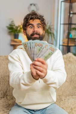 Başarılı genç Hintli adam elinde tuttuğu parayla evdeki kanepede oturuyor. Mutlu yetişkin Hindu adam çevrimiçi piyango oyunu kazanarak tatil planlama bütçe hesaplaması yapıyor..