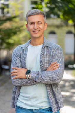 Kollarını kavuşturup gülümseyen mutlu olgun bir adamın portresi, kameranın rahatlamasına bakan mutlu yüz ifadesi dışarıda iyi haberler veriyor. Şehir sokağındaki beyaz adam. Dikey.