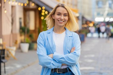 Mutlu, genç, sarışın, güler yüzlü, mutlu bir kadının portresi. Kameraya bakıyor, dinlenme hayali kuruyor, dışarıda iyi haberlerden tatmin oluyor. Şehir caddesinde duran kadın. Kasaba yaşam tarzları
