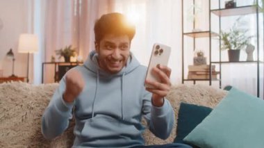 Online oyun kazananı ya da iyi haberi görünce şaşıran akıllı telefon tutan heyecanlı Hintli genç adam. Koltukta oturan mutlu adam başarının sevinci içinde kollarını muzaffer bir şekilde kaldırıyor, oturma odasında zaferi kutluyor..