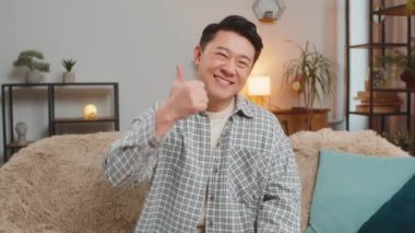 Asyalı bir adam evinde kanepede oturmuş mutlu ve tatmin olmuş hissediyordu, ışıl ışıl gülümsüyordu ve başparmaklarını hareket gibi kaldırıyordu. Oturma odasında rahat bir şekilde. Onaylama, neşe ve olumlu duygular.
