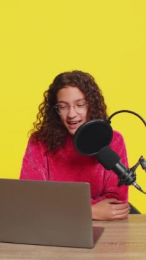 Masa başında podcast yapan, atölye veren, Webinar kullanan mutlu blogger kızı. Genç çocuk nüfuzlu mikrofon ve dizüstü bilgisayara konuşan webcam sarı arka planda görüntülü konuşma yapıyor.