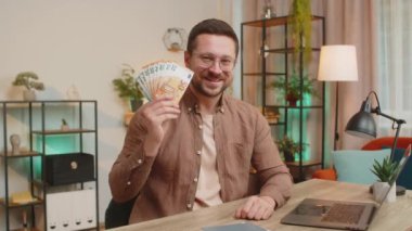 Genç işadamı elinde nakit para banknotlarının hayranı, başparmağıyla işaret ediyor, kutluyor, başarı kariyeri, çevrimiçi gelir, ev ofisinin oturma odasındaki zenginlik..