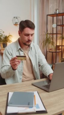Merkez ofisteki online alışveriş ödemeleri için banka kartı ve dizüstü bilgisayar kullanan Kafkasyalı mutlu bir serbest çalışan. İş anlaşması. Gülümseyen adam, bilgisayar E-Bankacılık uygulama servisiyle alışveriş yapıyor. Dikey.