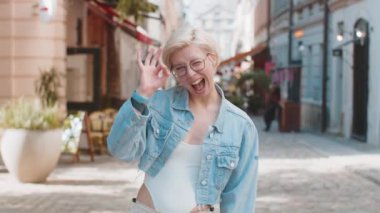 Çekici genç milenyum kızının portresi el işaretini gösteren kameraya bakıyor. Kısa sarı saçlı beyaz kadın her şeyi gösteriyor. Güzel başarı jesti vücut dili. Kameraya gülümseyen kadın.