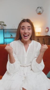 Mutlu genç bir kadın TV izlerken bağırıyor, başarıyı kutluyor, kazanıyor, gol başarısı iyi haber, piyango şansı, oturma odasında koltukta oturan zafer. Evde beyaz elbiseli bir kız var. Dikey