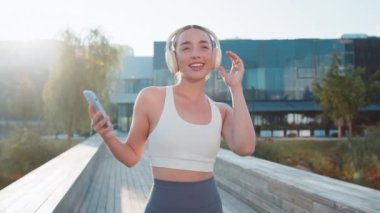 Genç spor koşucusu kadın eğitiminin portresi, kulaklık takmak, akıllı telefondan müzik dinlemek ve gülümsemek. Spor giyim antrenmanında sağlıklı bir kız sabah şehir parkında kardiyo yapıyor..