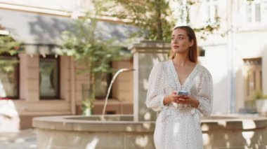 Gezgin genç bir kadın akıllı telefon kullanıyor cep telefonu navigatör uygulamasında haritada arama yapmak için kısa mesajlar yazıyor. Beyaz elbiseli kız şehir sokağının önünden geçiyor..