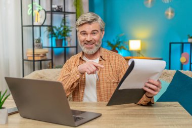 Beyaz, olgun bir adam evde oturmuş önemli belgeleri inceliyor ve dizüstü bilgisayar kullanıyor. Orta yaşlı bir adam başarılı bir işten aldığı memnuniyetle gülümser, verimlilik ve başarıyı gösterir.