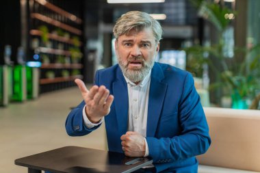 Çatışma Anlaşması 'nda. Olgun kızgın patron iş adamı webcam çevrimiçi video görüşmesi yapıyor, kameraya bakarak iş arkadaşlarıyla konuşuyor. Takım elbiseli beyaz adam modern ofis lobisinde oturuyor..