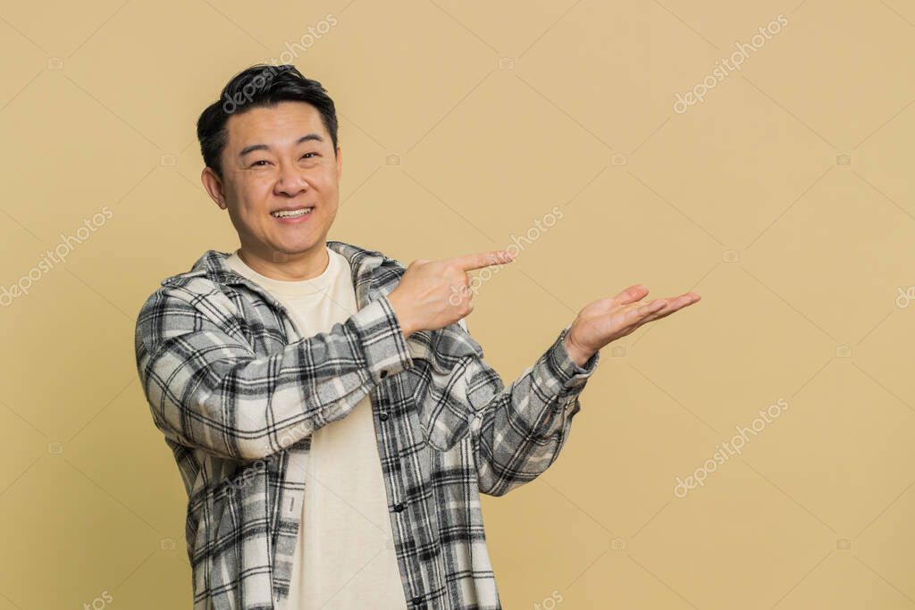 Asian middle-aged man showing thumbs up and pointing right empty place, advertising area for commercial discount text, copy space for goods promotion advertisement. Chinese guy on beige background