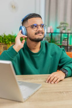 Happy relaxed Indian man in wireless headphones dancing at home office table workplace choosing listening favorite energetic disco rock n roll music entertaining. People weekend activities. Vertical clipart