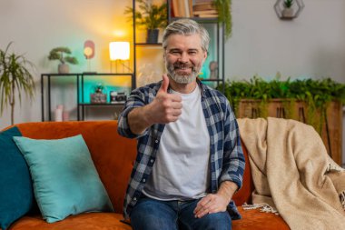 Mutlu, heyecanlı, Kafkas kıdemli bir adam kameraya bakıyor ve olumlu haber olumlu geri bildirim gibi başparmaklarını kaldırıyor. Oturma odasındaki koltukta oturan yaşlı sakallı, olgun bir adam..